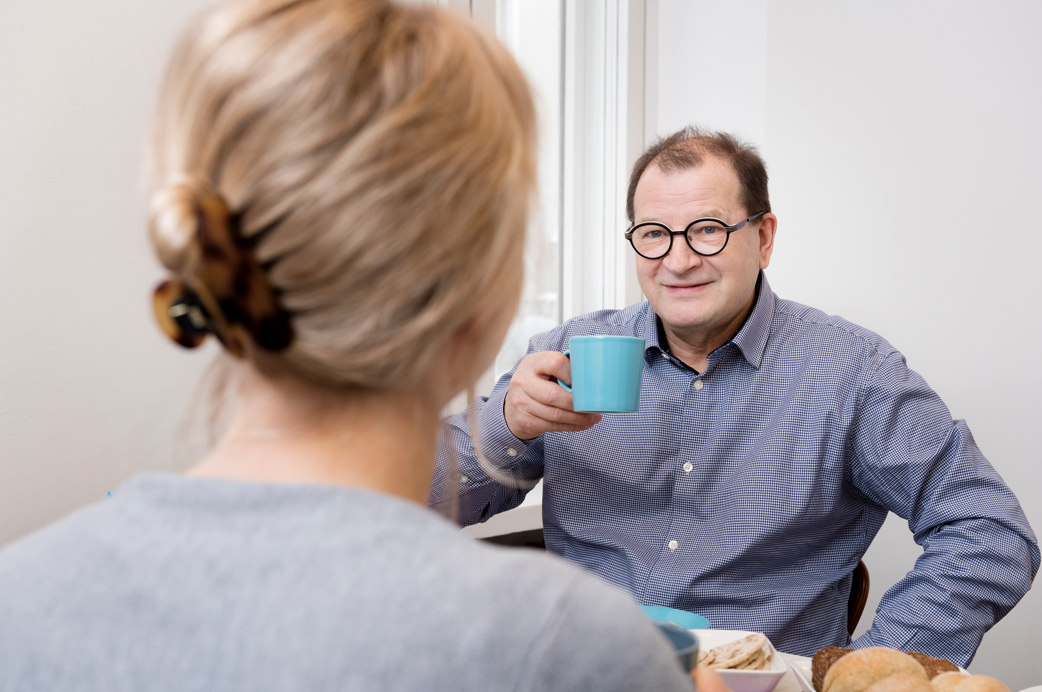 Mies pitää kädessään kahvikuppia, katsoo hymyillen edessään istuvaa naista..
