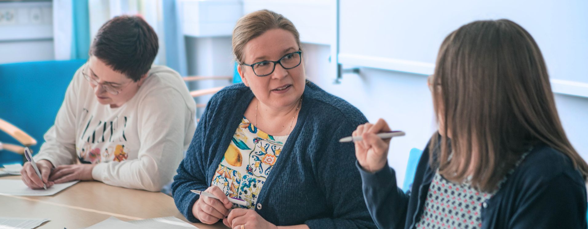 Kolme ihmistä työskentelee pöydän ääressä. Kaksi keskustelee ja yksi kirjoittaa.