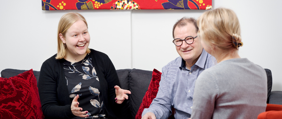 Kolme henkilöä istuu pöydän äärellä, yksi selkä kohti kameraa. Kaksi henkilöä istuu vierekkäin, hymyillen.