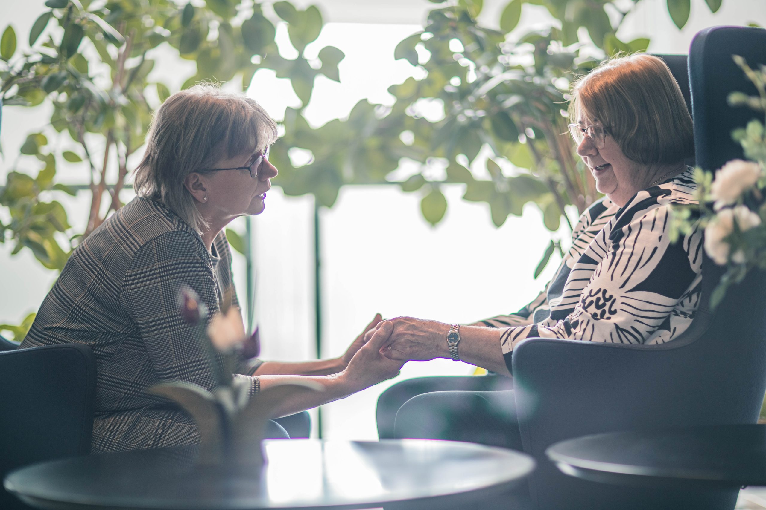 Nainen istuu penkillä ja pitelee käsissään toisen naisen käsiä, joka istuu nojatuolissa. Tunnelma on empaattinen.