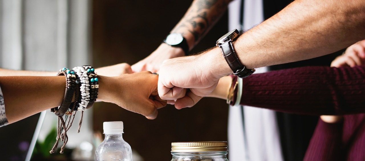 Joukko nuoria aikuisia tekee toisilleen rystysillä tervehdyksen. Alla pöytä, jossa tavaroita, mm. uomapullo ja lasipurkki, jossa pähkinöitä ja hedelmiä.